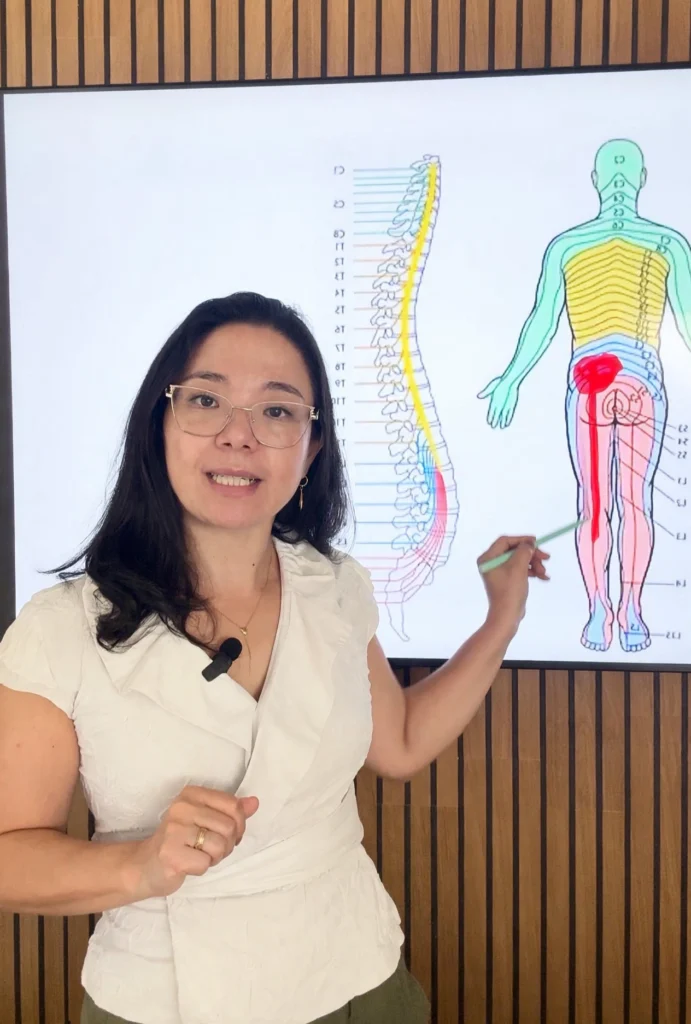 Dra Cris Nagae fisioterapeuta especializada no tratamento de hernia de disco em pe em frente a tela mostrando o caminho da dor irradiada da coluna ao longo da perna